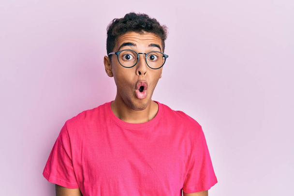 Молодий афроамериканець, одягнений в окуляри над рожевим фоном, наляканий і шокований несподіваним виразом, страхом і збудженим обличчям..  - Фото, зображення