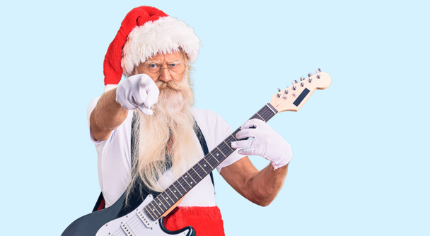 Old senior man with grey hair and long beard wearing santa claus costume playing electric guitar pointing with finger to the camera and to you, confident gesture looking serious  - Photo, Image