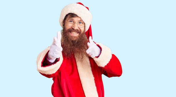 Uzun sakallı, Noel Baba kostümü giyen yakışıklı kızıl saçlı adam mutlu ve komik yüzlü parmakları kameraya doğrultuyor. iyi enerji ve titreşimler.  - Fotoğraf, Görsel