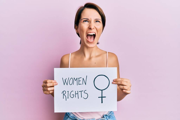 Young brunette woman with short hair holding woman rights banner angry and mad screaming frustrated and furious, shouting with anger looking up.  - Foto, Bild