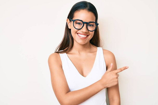 Mladá krásná latinská dívka na sobě ležérní oblečení a brýle s veselým úsměvem ukazuje rukou a prstem nahoru  - Fotografie, Obrázek