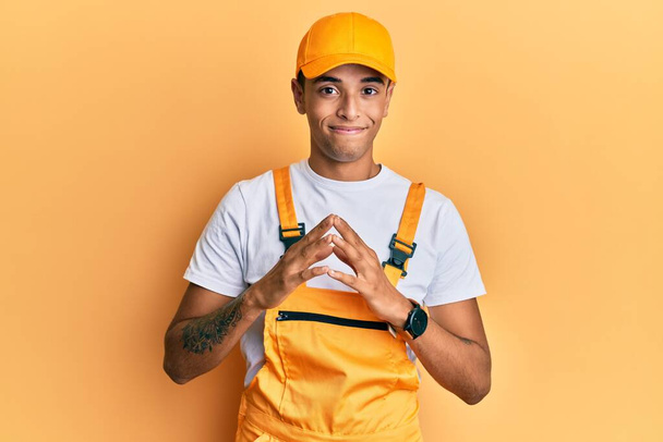 Young handsome african american man wearing handyman uniform over yellow background hands together and fingers crossed smiling relaxed and cheerful. success and optimistic  - Photo, Image