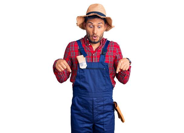 Handsome latin american young man weaing handyman uniform pointing down with fingers showing advertisement, surprised face and open mouth  - 写真・画像