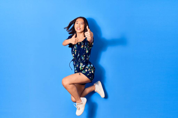 Young beautiful chinese woman wearing casual clothes smiling happy. Jumping with smile on face doing ok sign with thumbs up over isolated blue background - Foto, Imagen