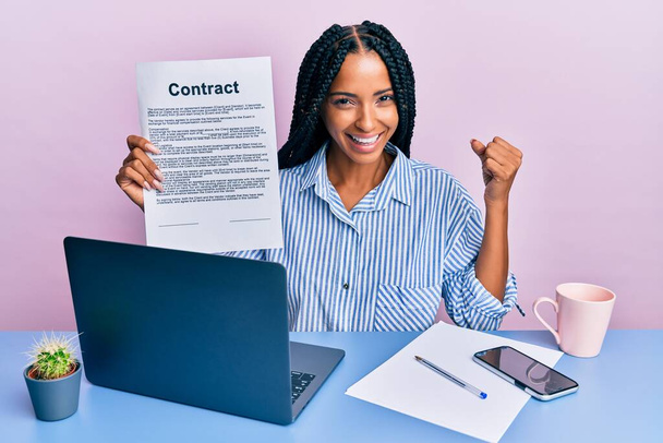 Beautiful hispanic woman at the office showing contract document screaming proud, celebrating victory and success very excited with raised arm  - Foto, immagini
