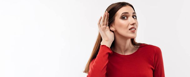 Jonge mooie brunette vrouw in casual rode trui hand in hand in de buurt van oor proberen om iets te luisteren. Studio opname, witte achtergrond. Menselijke emoties, gezichtsuitdrukking concept - Foto, afbeelding