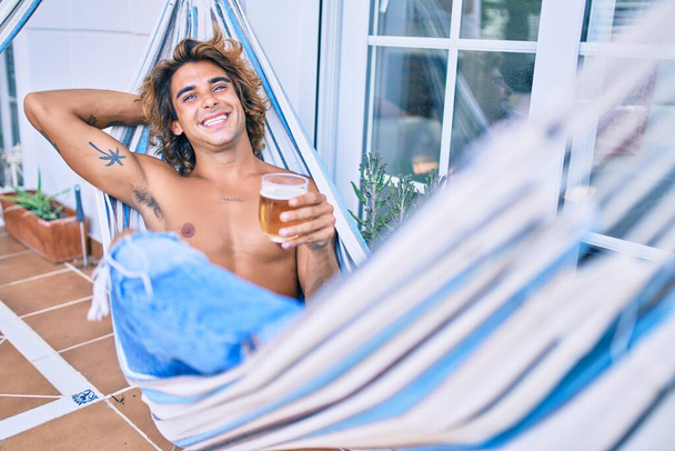Giovane uomo ispanico rilassato bere birra sdraiato sull'amaca in terrazza. - Foto, immagini