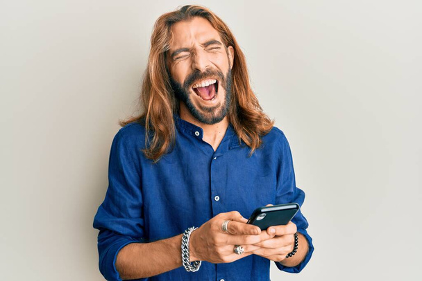 Ελκυστικός άνθρωπος με μακριά μαλλιά και γενειάδα χρησιμοποιώντας smartphone χαμογελώντας και γελώντας δυνατά γιατί αστείο τρελό αστείο.  - Φωτογραφία, εικόνα