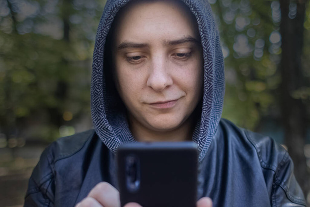 Concept. A man in a leather jacket with a hood looks at the phone and smiles like a maniac or a telephone scammer. Theft of information from a smartphone. Correspondence of a maniac on social networks - Photo, Image