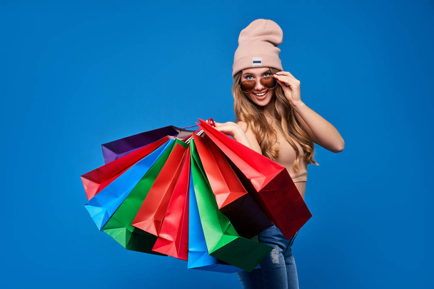 Photo of a beautiful young blonde woman in sunglasses and a hat with packages on sale. Happy girl after shopping online. - Zdjęcie, obraz