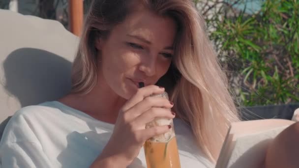 Medium closeup of young model sipping mixed drink and reading paper book smiling in posh resort terrace - Footage, Video