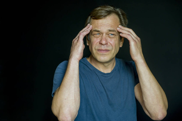 Porträt eines 45-50-jährigen Mannes, der seinen Kopf in den Händen hält, vor dunklem Hintergrund. Er kann häufige Kopfschmerzen oder Migräne haben. - Foto, Bild
