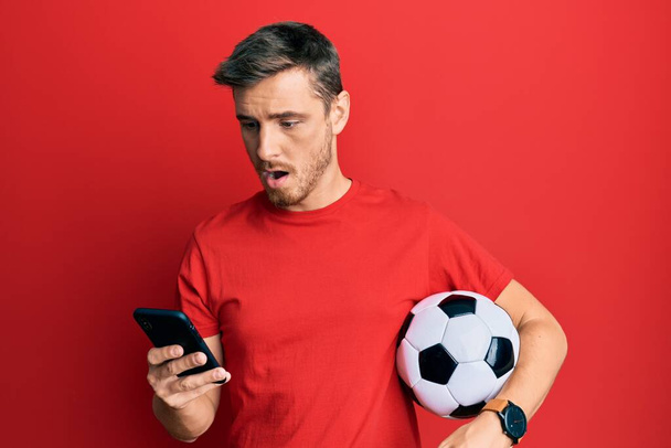 Handsome caucasian man holding football ball looking at smartphone afraid and shocked with surprise and amazed expression, fear and excited face.  - Photo, Image
