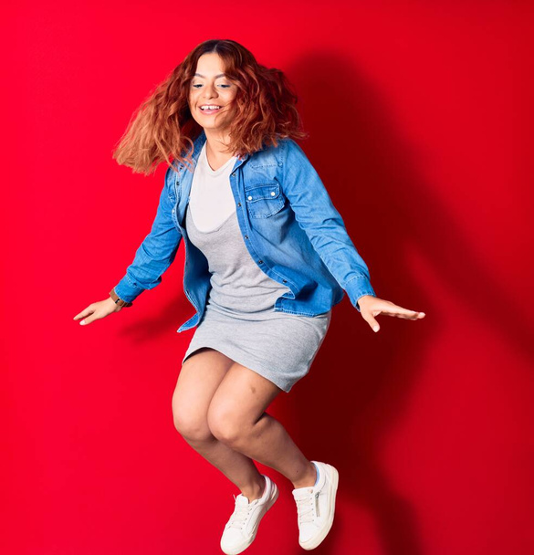 Young beautiful latin woman wearing casual clothes smiling happy. Jumping with smile on face over isolated red background - Zdjęcie, obraz