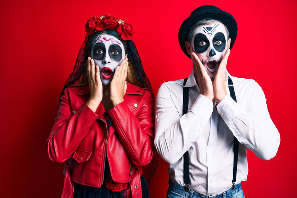 Couple wearing day of the dead costume over red afraid and shocked, surprise and amazed expression with hands on face  - Photo, Image