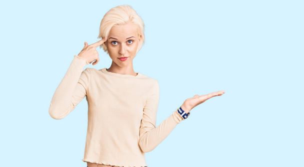 Jeune femme blonde portant des vêtements décontractés confus et agacés avec la paume ouverte montrant l'espace de copie et pointant du doigt vers le front. Penses-y..  - Photo, image