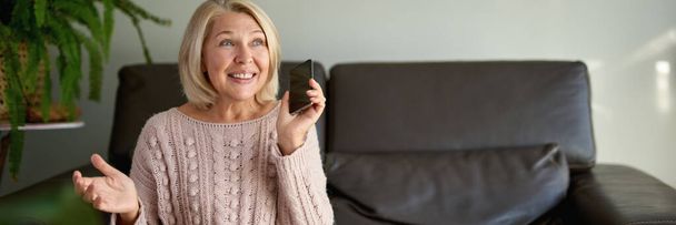 Mujer mayor usando el teléfono móvil mientras está sentada en el sofá. - Foto, Imagen