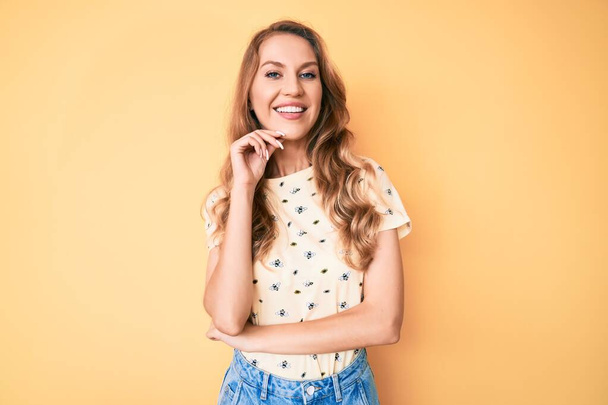 Young caucasian woman with blond hair wearing casual summer clothes smiling looking confident at the camera with crossed arms and hand on chin. thinking positive.  - Fotografie, Obrázek