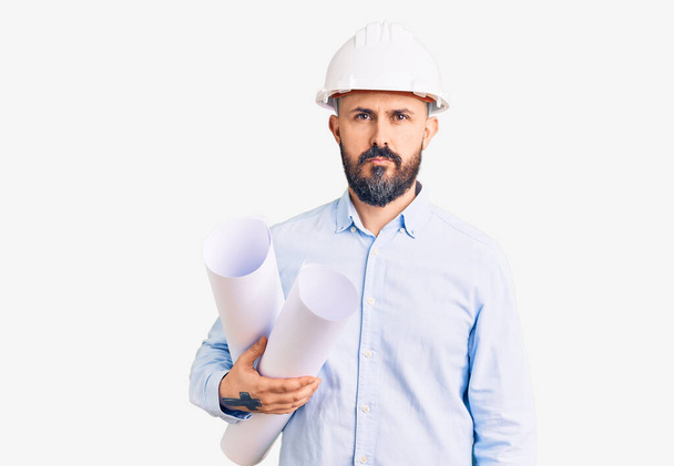Young handsome man wearing hardhat holding paper blueprints thinking attitude and sober expression looking self confident  - Photo, image