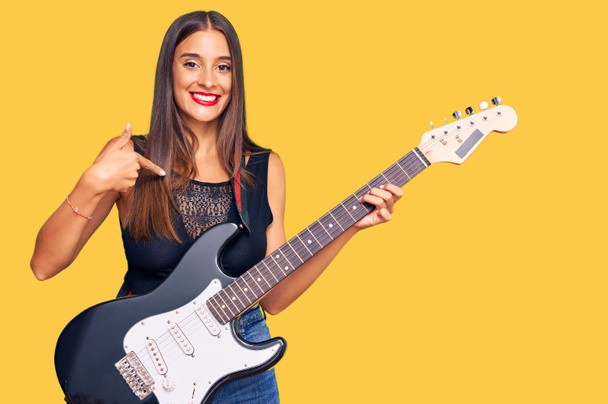 Молода іспаномовна жінка грає на електрогітарі, вказуючи пальцем на себе, усміхаючись щасливою і гордою
  - Фото, зображення