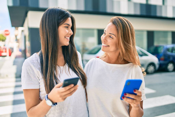 Schöne hispanische Mutter und Tochter lächeln glücklich per Smartphone in die Stadt. - Foto, Bild