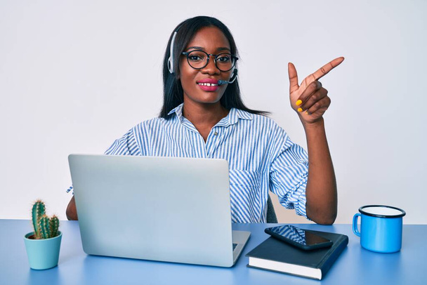 Молодая африканская американка, работающая в офисе, в наушниках оператора, улыбающаяся, счастливая, указывая рукой и пальцем в сторону.  - Фото, изображение