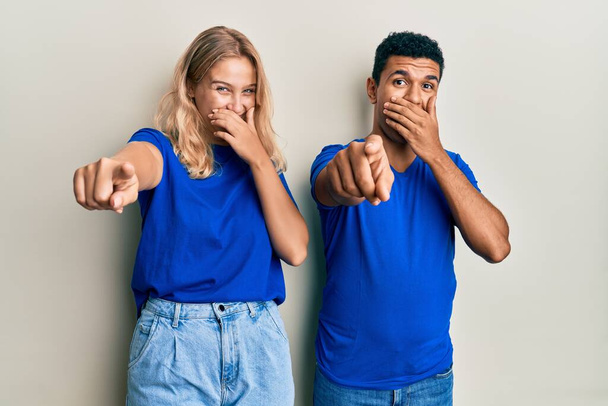 Junge interrassische Paare in lässiger Kleidung lachen dich an, zeigen mit der Hand über den Mund in die Kamera, schämen sich  - Foto, Bild