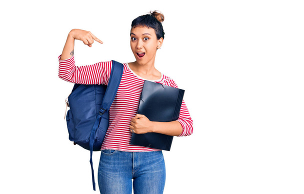 Junge Frau im Studentenrucksack hält Ordner und lächelt glücklich und zeigt mit Hand und Finger  - Foto, Bild