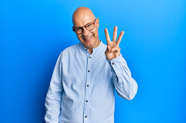 Mann mittleren Alters mit Glatze trägt lässige Kleidung und Brille und zeigt mit Fingern Nummer drei nach oben, während er selbstbewusst und glücklich lächelt.  - Foto, Bild