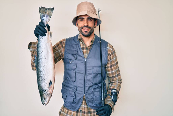 Handsome hispanic man with beard holding fishing rod and raw salmon smiling with a happy and cool smile on face. showing teeth.  - Fotó, kép