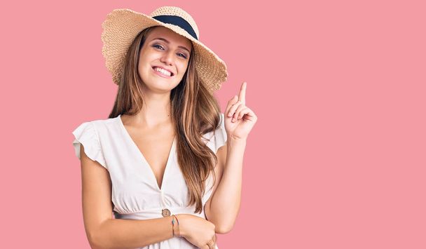 Jeune belle femme blonde portant une robe d'été et un chapeau avec un grand sourire sur le visage, pointant avec la main et le doigt vers le côté en regardant la caméra.  - Photo, image
