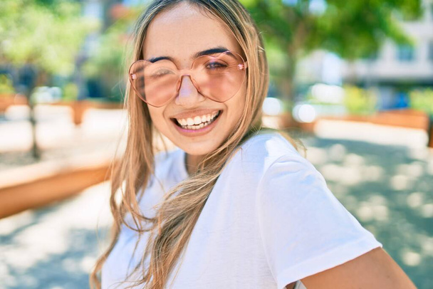 Young beautiful blonde caucasian woman smiling happy outdoors on a sunny day wearing heart shaped sunglasses - Photo, Image
