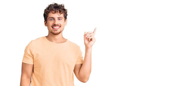 Νεαρός όμορφος άντρας με σγουρά μαλλιά φορώντας καθημερινά ρούχα χαμογελώντας χαρούμενος δείχνοντας με το χέρι και το δάχτυλο στο πλάι  - Φωτογραφία, εικόνα