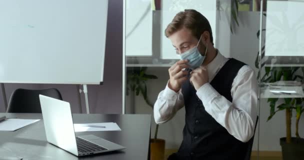 Businessman freelancer in face medical mask sitting at table messaging online on Covid-19 lockdown. Caucasian man typing on laptop keyboard. Manager working remote from office in coronavirus pandemic - Πλάνα, βίντεο