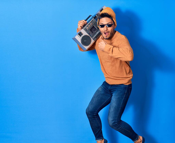 Young handsome latin man smiling happy wearing thug lif sunglasses. Jumping with smile on face holding boombox doing ok sign with thumb up over isolated blue background - Foto, afbeelding