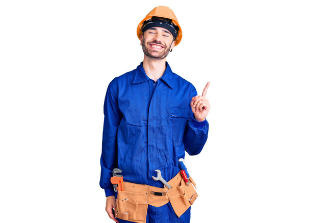 Joven hispano vistiendo uniforme de trabajador con una gran sonrisa en la cara, señalando con el dedo de la mano hacia un lado mirando a la cámara.  - Foto, Imagen