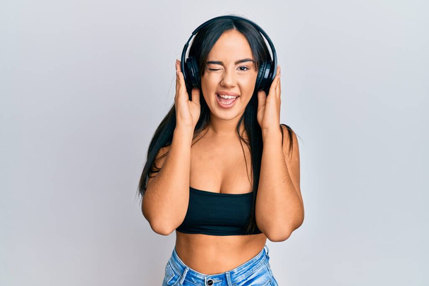 Νέα όμορφη ισπανόφωνη κοπέλα ακούγοντας μουσική χρησιμοποιώντας ακουστικά να κλείνει το μάτι κοιτάζοντας την κάμερα με σέξι έκφραση, χαρούμενο και χαρούμενο πρόσωπο.  - Φωτογραφία, εικόνα