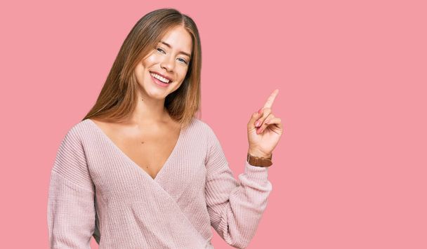 Krásná blondýnka na sobě ležérní zimní růžový svetr s velkým úsměvem na tváři, ukazující ukazováčkem na stranu při pohledu do kamery.  - Fotografie, Obrázek