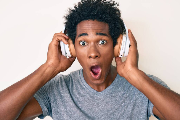 Handsome african american man with afro hair listening to music using headphones afraid and shocked with surprise and amazed expression, fear and excited face.  - Photo, image