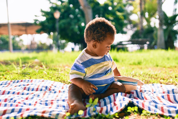 Adorable african american toddler holding hat sitting on the grass at the park. - Foto, Imagem