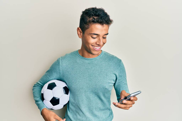 Junger gutaussehender afrikanisch-amerikanischer Mann mit Fußballball, der mit einem fröhlichen und kühlen Lächeln auf dem Gesicht auf sein Smartphone blickt. Zähne zeigen.  - Foto, Bild