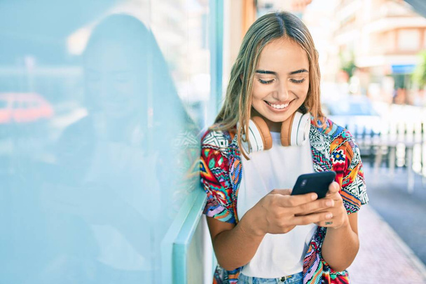 Young beautiful blonde caucasian woman smiling happy outdoors on a sunny day wearing headphones and using smartphone - Photo, Image