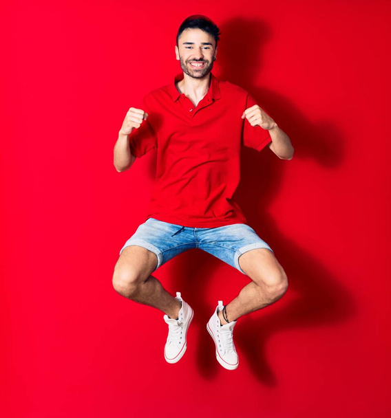 Young handsome deliveryman wearing cap smiling happy. Jumping with smile on face doing winner gesture with fists up over isolated red background - Photo, Image