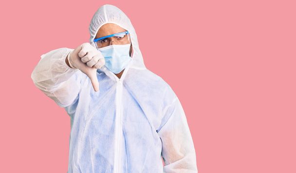 Jonge Spaanse man draagt doktersbescherming coronavirus uniform en medisch masker kijken ongelukkig en boos tonen afwijzing en negatief met duimen naar beneden gebaar. slechte uitdrukking.  - Foto, afbeelding