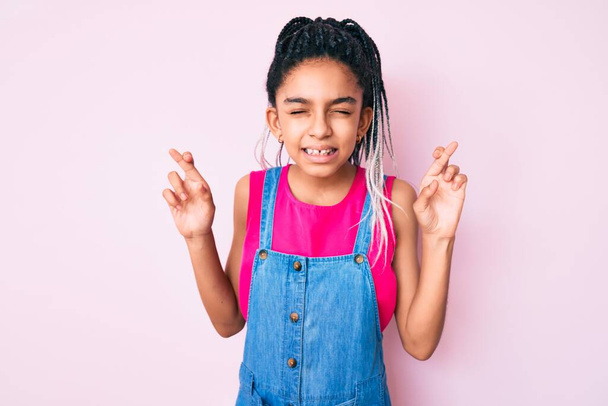 Young african american girl child with braids wearing casual clothes over pink background gesturing finger crossed smiling with hope and eyes closed. luck and superstitious concept.  - Photo, Image