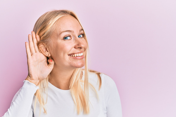 Молода блондинка в повсякденному одязі посміхається рукою через слух слухаючи чутки або плітки. концепція глухоти
.  - Фото, зображення