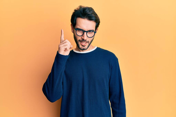 Junger hispanischer Mann in lässiger Kleidung und Brille mit erhobenem Zeigefinger und erfolgreicher Idee. aufgeregt und glücklich. Nummer eins.  - Foto, Bild