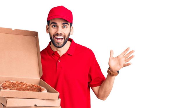 Junger gutaussehender Mann mit Bart trägt Lieferuniform und hält Schachteln mit Pizza in der Hand und feiert den Sieg mit glücklichem Lächeln und Siegerausdruck mit erhobenen Händen  - Foto, Bild
