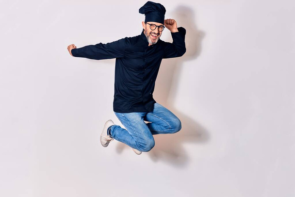 Middle age handsome hispanic man wearing glasses and cook uniform smiling happy. Jumping with smile on face celebrating with fists up over isolated white background - Foto, Imagen