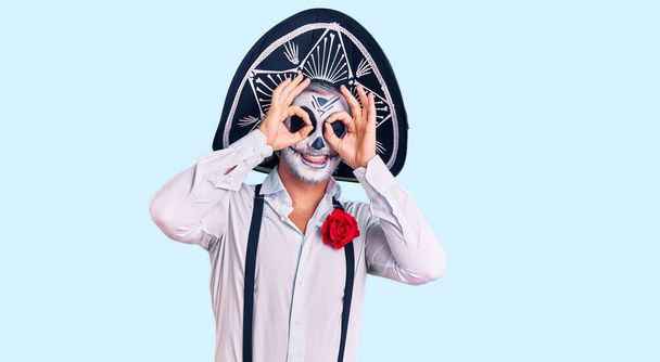 Man wearing day of the dead costume over background doing ok gesture like binoculars sticking tongue out, eyes looking through fingers. crazy expression.  - Photo, Image